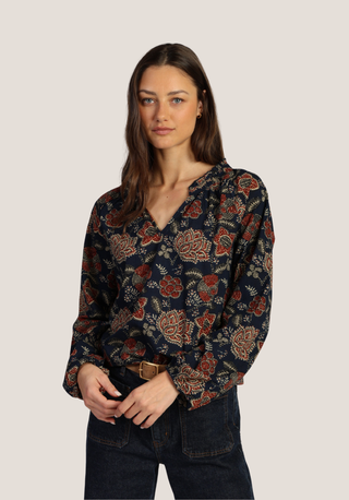 A woman modeling a dark navy floral blouse with red, beige, and green botanical patterns, paired with dark blue jeans, against a solid light gray background.