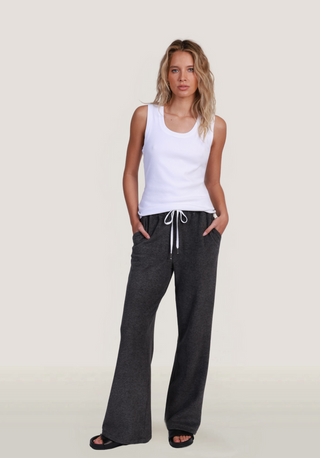 A woman modeling dark charcoal gray wide-leg lounge pants with a white drawstring, paired with a white tank top and black sandals, against a light beige studio background.