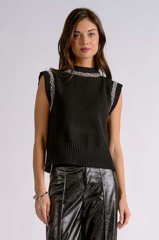 A woman modeling a sleeveless black knit top with silver rhinestone trim at the mock neck and along the cap sleeves, paired with shiny black pants, against a light gray studio background.