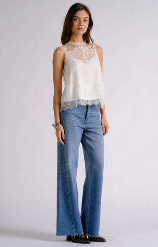 A model stands against a light gray studio background wearing a white sleeveless lace-trim camisole top and wide-leg blue jeans.