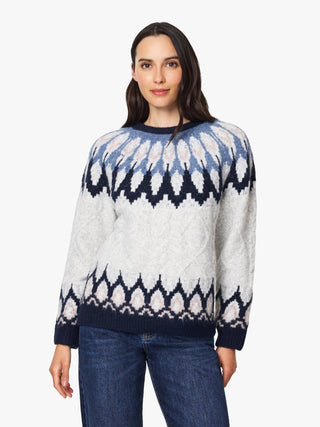 A woman modeling a long-sleeve knit sweater with a geometric Fair Isle pattern in navy blue, light blue, and off-white, paired with blue jeans against a plain white background.