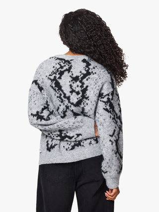 A person shown from the back wearing a light gray knit sweater with a black abstract pattern and black pants, against a plain light background.