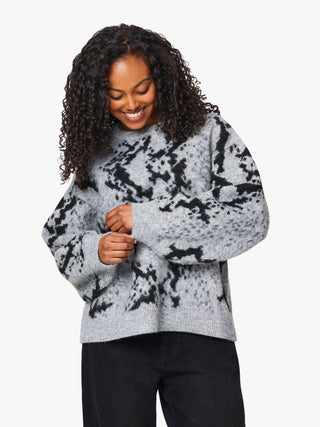 A woman with curly black hair wearing a light gray knit sweater with a black mottled or tie-dye pattern, paired with black pants, against a plain light gray background.