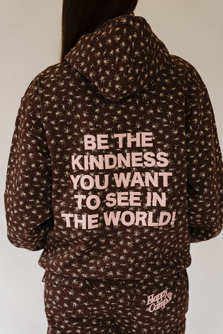 A person shown from the back wearing a dark brown patterned hoodie with light pink text that reads 'BE THE KINDNESS YOU WANT TO SEE IN THE WORLD!' and matching bottoms. The hoodie has a small repeating floral or star-like print and a visible 'Happy Camp3r' logo near the lower right leg.