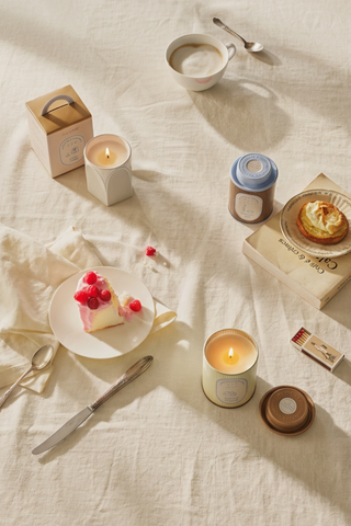 A set table with lit candles from Illume, surrounded by various desserts and a cup of tea, with a book in the background.