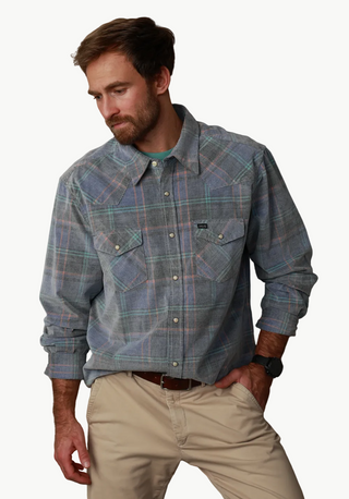 A man wearing a blue and gray plaid western-style button-down shirt with beige pants, leaning against a textured gray wall.
