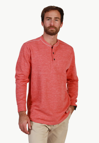 A man wearing a coral red heathered long-sleeve henley shirt and beige pants, standing against a textured light gray wall.