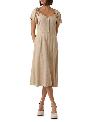 A woman standing, wearing a beige dress with white polka dots and a round neckline.