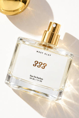 A clear glass perfume bottle with a white label and gold spray top, photographed on a bright white background with strong sunlight and shadows. The label reads 'WEST CLAY' and '333 Eau De Parfum 50 ML / 1.