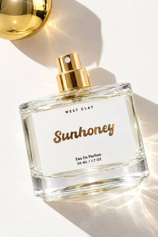 A 50 ml glass perfume bottle labeled 'Sunhoney' by West Clay, featuring a gold spray nozzle and a white label with gold lettering, set against a bright white background with dramatic golden light rays and a matching gold sphere prop.