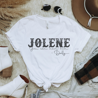 A white graphic t-shirt with the text 'JOLENE you can have him - Dolly' printed in black, styled in a flat lay with sunglasses, a hat, white boots, and a plaid shirt on a faux fur background.