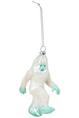A hanging Christmas ornament shaped like a white yeti with mint green accents on its face, hands, and feet, suspended from a silver string against a plain white background.